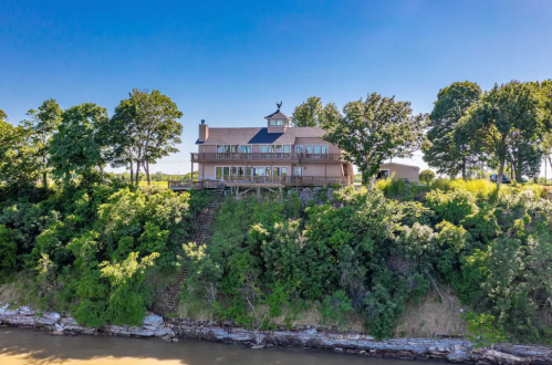A large house on a hillside overlooking a river, surrounded by trees under a clear blue sky.