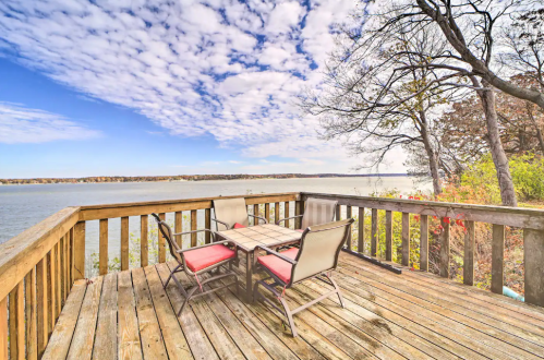 A wooden deck overlooking a calm lake, featuring a table and chairs under a partly cloudy sky.