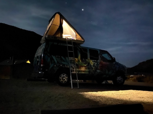 A colorful van with a rooftop tent, parked at night under a starry sky, illuminated by a nearby light.