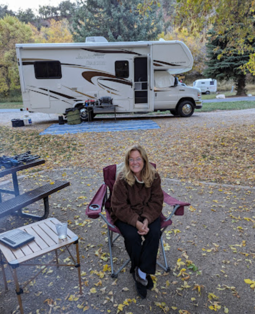 A woman sits in a camping chair outside an RV, surrounded by autumn leaves and a picnic setup.