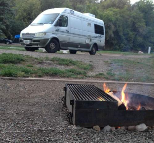A camper van parked near a fire pit with flames, surrounded by greenery and gravel.