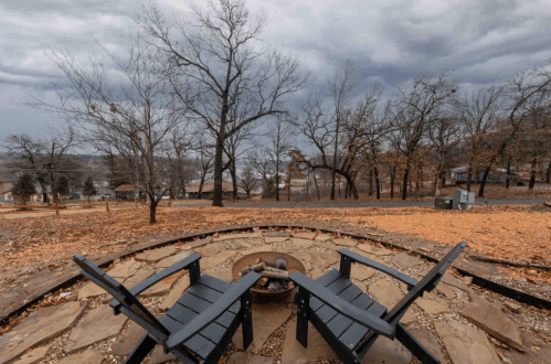 Two black chairs facing a fire pit, surrounded by a stone circle, with a cloudy sky and bare trees in the background.