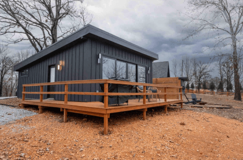 A modern black cabin with a wooden deck, surrounded by trees and a cloudy sky.
