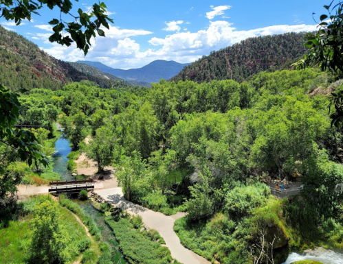Lush green landscape with a river, bridge, and mountains under a blue sky with scattered clouds.