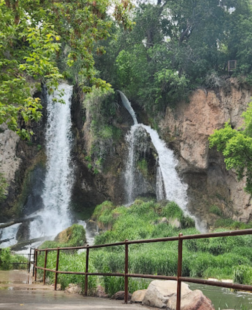 A scenic view of two waterfalls cascading over rocky cliffs, surrounded by lush greenery and a metal railing in the foreground.