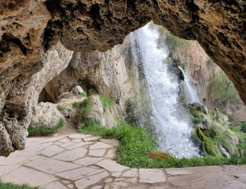 A waterfall cascades down rocky cliffs, viewed through a natural stone arch, with lush greenery surrounding the scene.