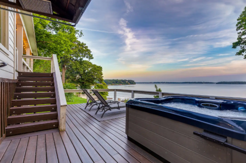 A scenic deck with a hot tub, lounge chair, and stairs overlooking a calm lake at sunset.