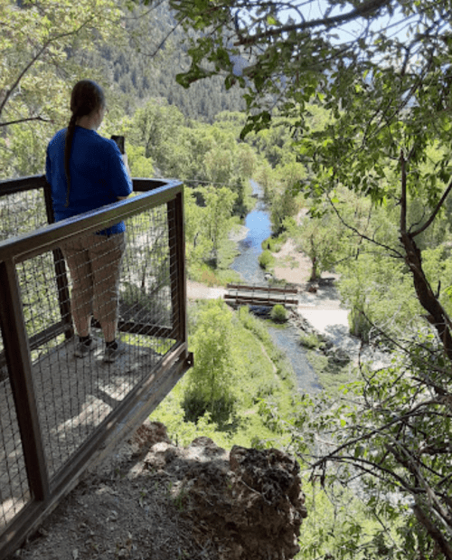 A person stands on a viewing platform overlooking a lush green landscape and a winding river below.