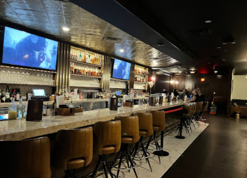 A stylish bar with a marble countertop, high stools, and two large screens displaying images. Dimly lit atmosphere.