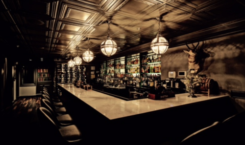 A dimly lit bar with a stylish design, featuring a long counter, shelves of bottles, and decorative lighting.
