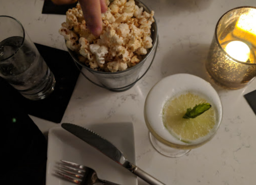 A table setting with a glass of cocktail, a bowl of popcorn, and a lit candle, creating a cozy atmosphere.