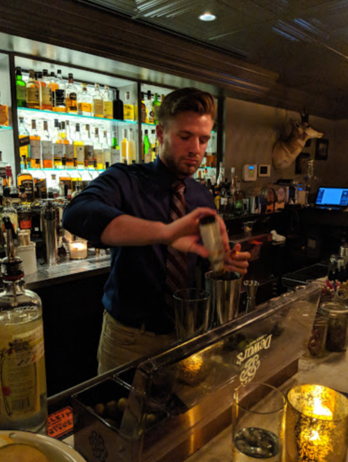 A bartender in a blue shirt and tie mixes a drink at a well-stocked bar with various bottles in the background.