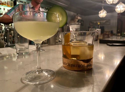A cocktail glass with a cucumber garnish and a rocks glass with a whiskey drink on a bar counter.