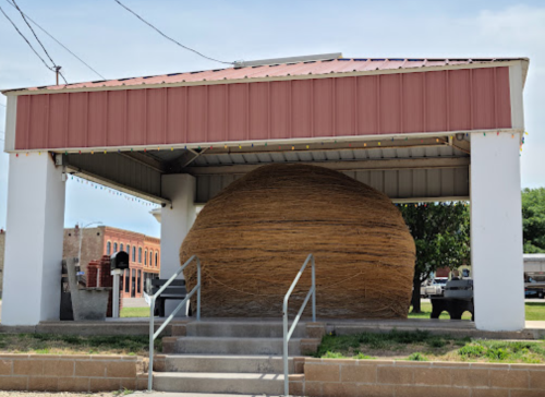 A large, round straw sculpture under a covered structure, with a grassy area and buildings in the background.