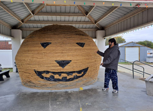 A person stands next to a large hay bale decorated with a smiling face, under a covered structure.