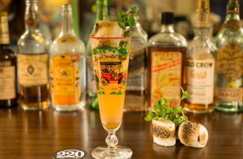 A cocktail in a tall glass garnished with herbs and a toasted marshmallow, with various liquor bottles in the background.