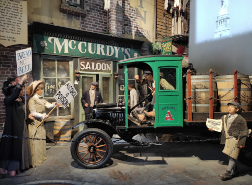 A vintage truck in a street scene with people holding signs outside a saloon, depicting a historical setting.