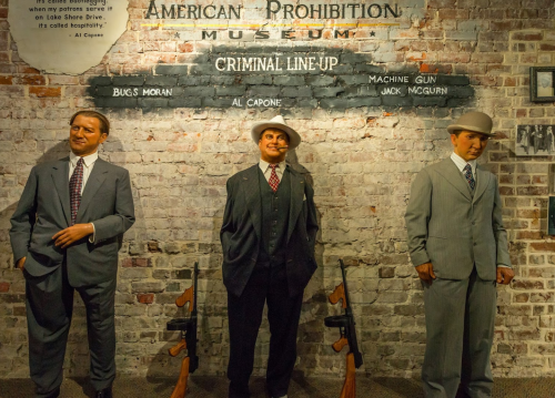 Three life-sized figures in suits stand in a criminal lineup at the American Prohibition Museum, flanked by vintage rifles.