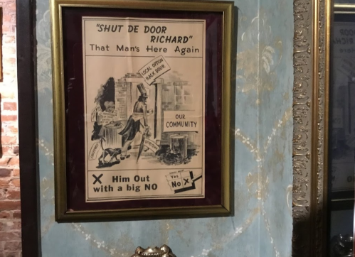 A vintage poster framed on a wall, urging to "Shut de Door Richard" with a humorous illustration and community message.