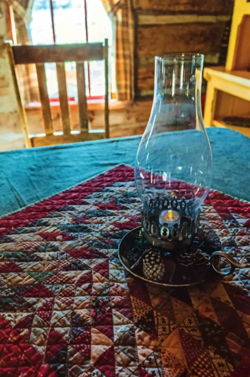 A glass oil lamp sits on a quilted tablecloth in a rustic room with wooden furniture and warm lighting.