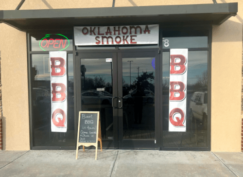 Exterior of "Oklahoma Smoke BBQ" restaurant with an open sign and banners promoting the best BBQ in town.