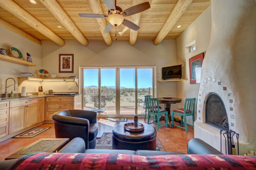 Cozy living space with wooden beams, a fireplace, kitchen area, and large windows showcasing a scenic view.
