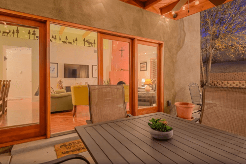Cozy outdoor seating area with a table, overlooking a warmly lit living room through large glass doors.