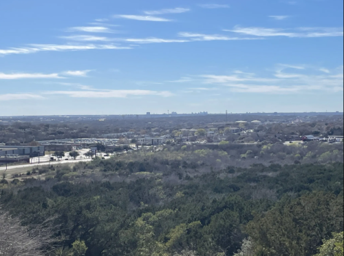 A panoramic view of a landscape with rolling hills, trees, and a distant city skyline under a clear blue sky.