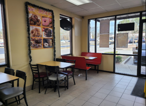 Interior of a donut shop with tables, red seating, and a wall display of donuts. Bright, inviting atmosphere.