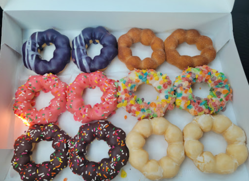 A box of colorful donuts in various flavors, including chocolate, pink, and purple, with sprinkles and cereal toppings.