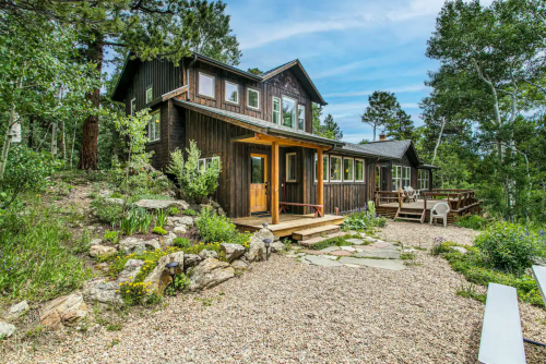 A rustic wooden house surrounded by greenery and rocks, featuring a porch and multiple windows under a blue sky.