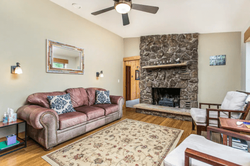 Cozy living room with a stone fireplace, brown sofa, wooden accents, and a decorative rug. Bright and inviting atmosphere.