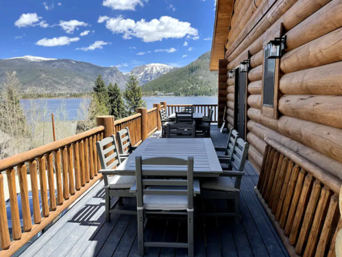 A wooden deck with a dining table and chairs overlooks a lake and mountains under a blue sky with fluffy clouds.