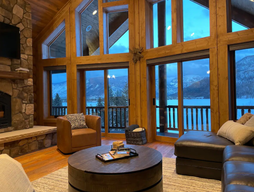 Cozy living room with large windows showcasing a snowy lake view, wooden beams, and a stone fireplace.