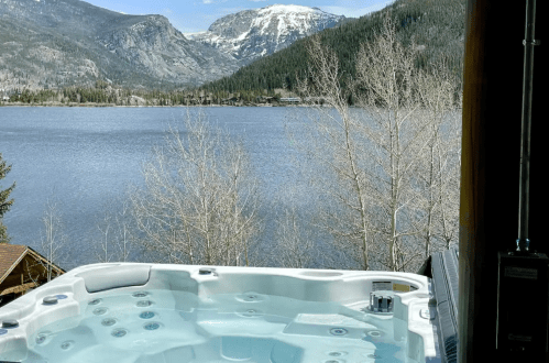 A hot tub overlooking a serene lake and snow-capped mountains, surrounded by trees and clear blue skies.