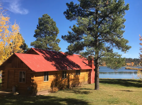 A rustic wooden cabin with a red roof, surrounded by trees, near a calm lake under a clear blue sky.