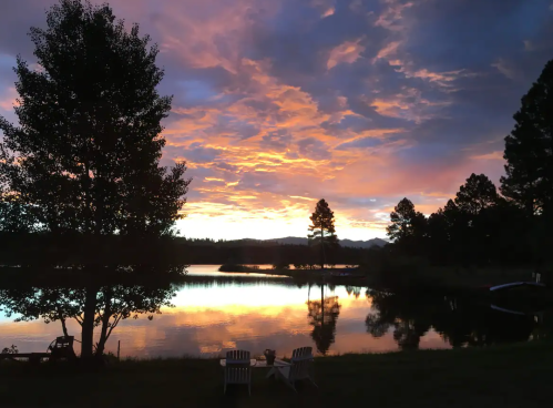 A serene lake at sunset, with colorful clouds reflecting on the water and two chairs by the shore.