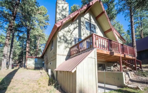A two-story cabin with a sloped roof, balcony, and surrounded by tall trees in a forested area.