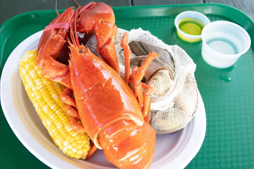 A plate with a cooked lobster, corn on the cob, and a bag of clams, accompanied by dipping sauces.