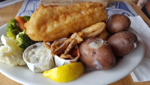 A plate with fried fish, calamari, boiled potatoes, steamed broccoli, and dipping sauces, garnished with a lemon wedge.