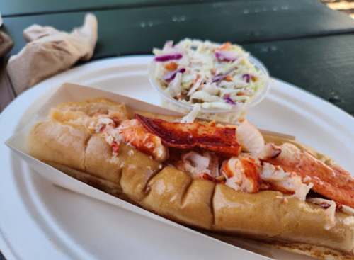 A lobster roll in a bun with a side of coleslaw on a plate, set on a green table.