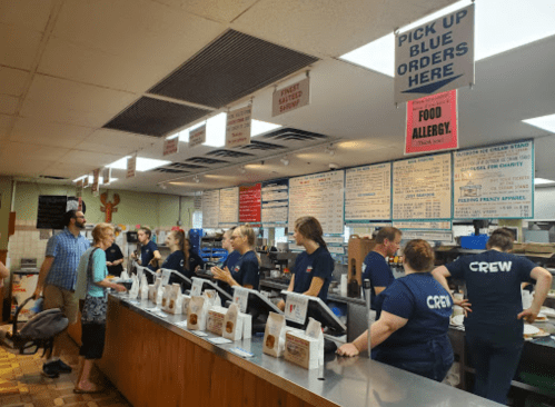 A busy restaurant counter with staff in blue shirts, customers ordering, and a menu displayed above. Signs for food allergies visible.