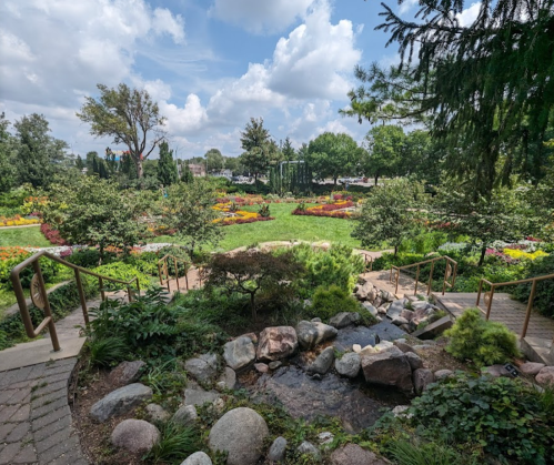 A lush garden with colorful flowers, greenery, and a small stream, under a partly cloudy sky.