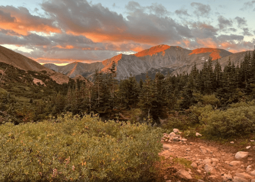 A scenic mountain landscape at sunset, with peaks illuminated by warm light and lush greenery in the foreground.