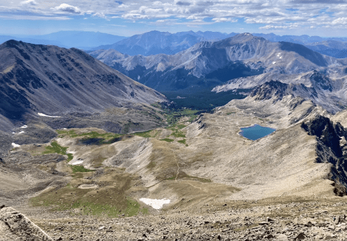 A panoramic view of rugged mountains and a serene blue lake nestled in a valley under a partly cloudy sky.