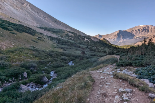 A scenic mountain landscape with a winding trail, lush greenery, and a stream flowing through the valley.