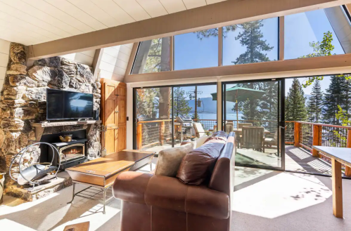 Cozy living room with stone fireplace, large windows, and a view of the lake and trees outside.