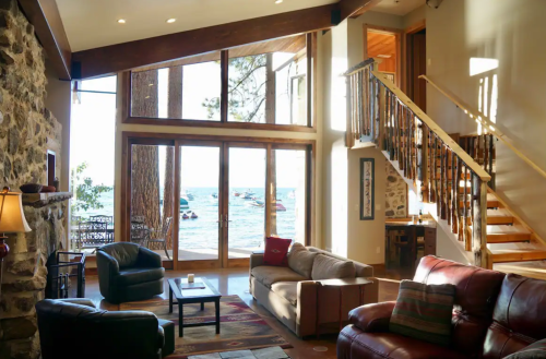 Cozy living room with large windows overlooking a lake, featuring a stone fireplace and comfortable seating.
