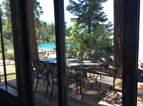 View of a lakeside deck with a dining table and chairs, surrounded by trees and a glimpse of people by the water.