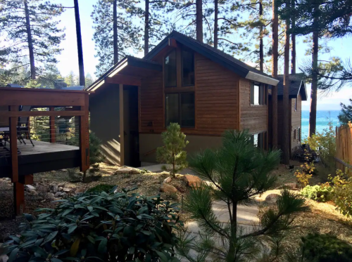 A modern wooden house surrounded by trees, with a pathway leading to a scenic view of a lake.